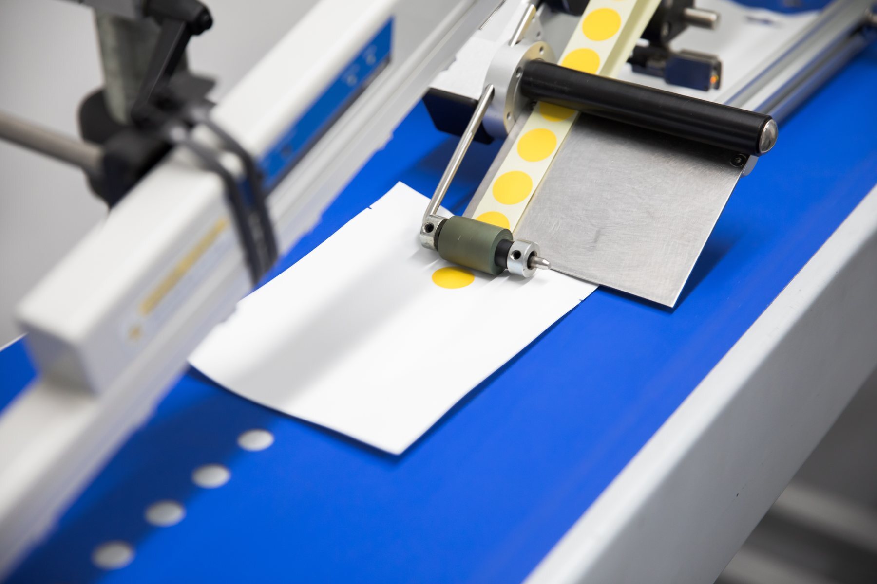 A single yellow gamma indicator dot being applied to a white card on a blue conveyor by the applicator roller, with the dot web feeding in above.