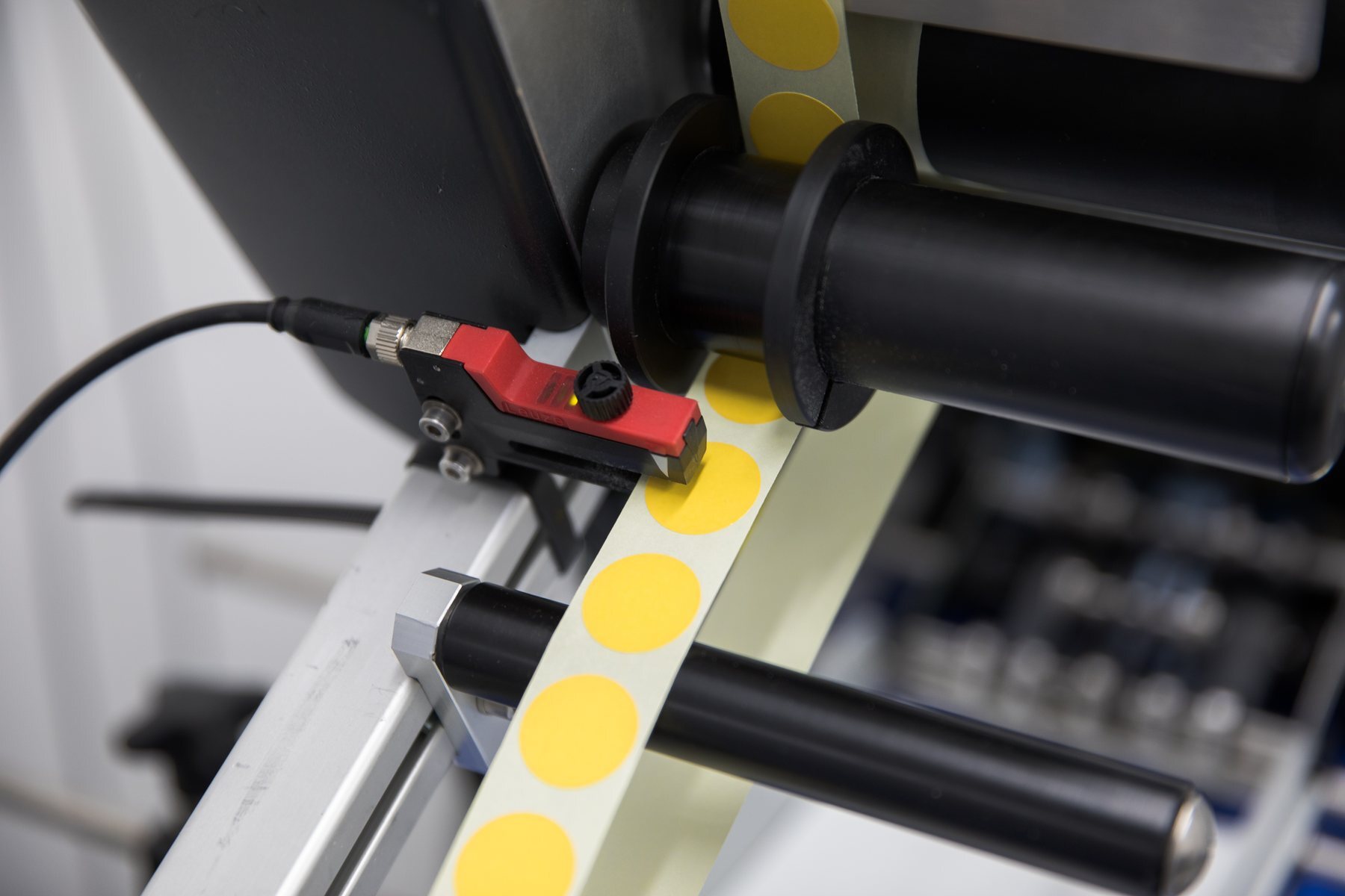 A web of yellow gamma indicator dots on backing liner running through a labelling machine, past a red sensor and over black rollers.
