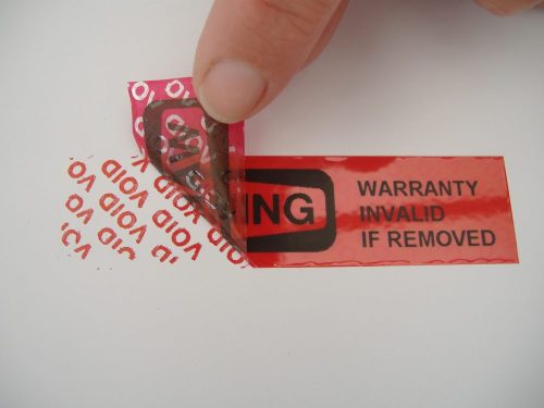A finger peeling back a red tamper-evident security label reading "WARRANTY INVALID IF REMOVED", exposing the red VOID VOID VOID residue pattern left behind on the surface.