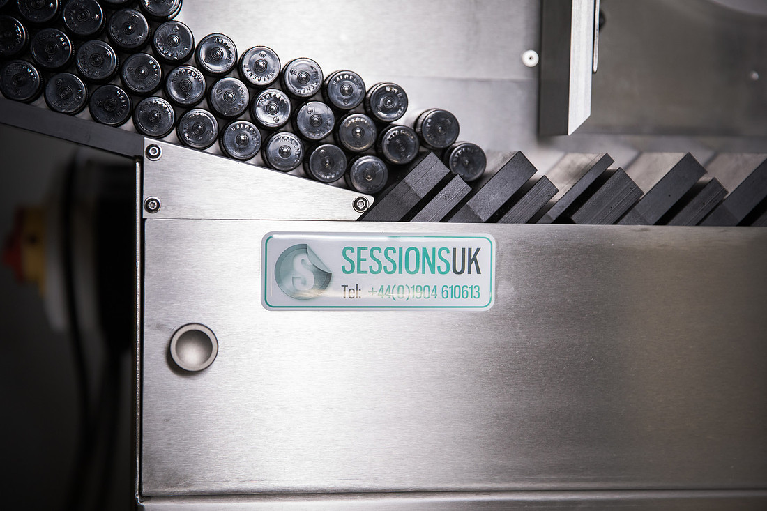 Black cylindrical canisters collected on a stainless steel machine chute, with a SESSIONSUK badge on the panel.