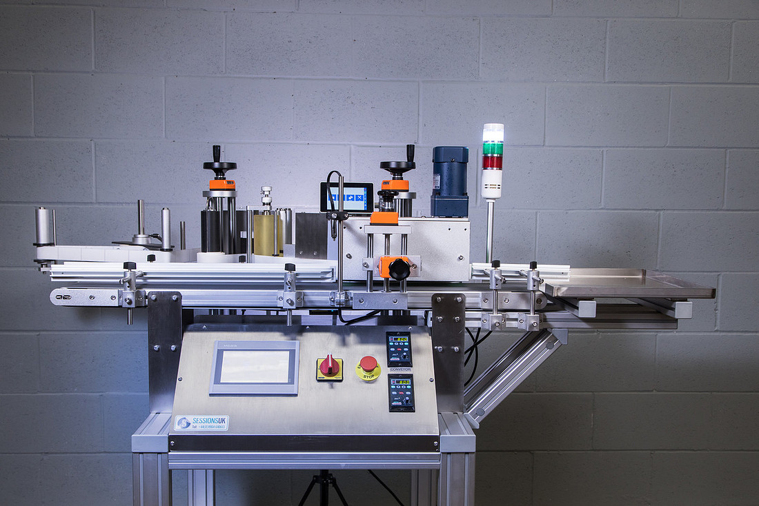 A bench-mounted inline labelling machine with conveyor, label reel and touchscreen control panel.