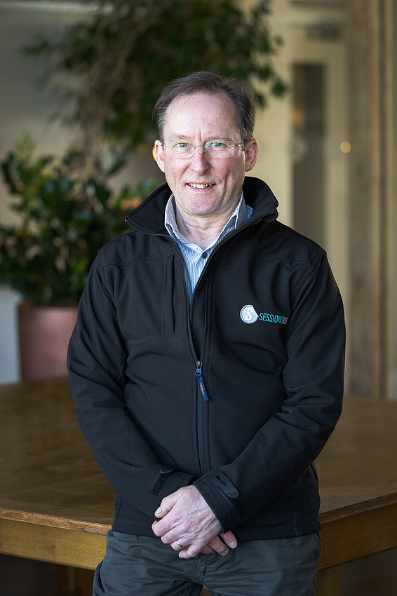 Portrait of a man wearing glasses and a navy jacket with the Sessions UK logo, standing indoors.