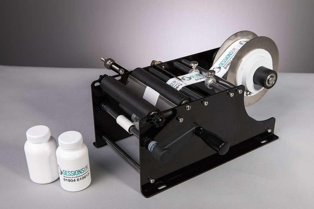 A black benchtop hand-crank wrap-round labeller on grey seamless with two white Sessions UK bottles beside it.