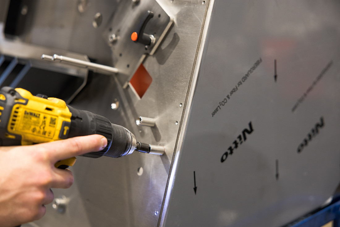 A hand and forearm driving a cordless drill into a stainless steel machine panel.