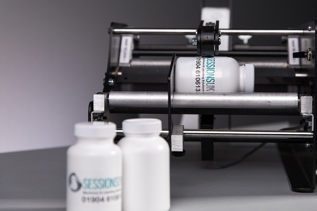 A black benchtop wrap-round labeller with a white bottle in the rollers and two labelled bottles in the foreground.