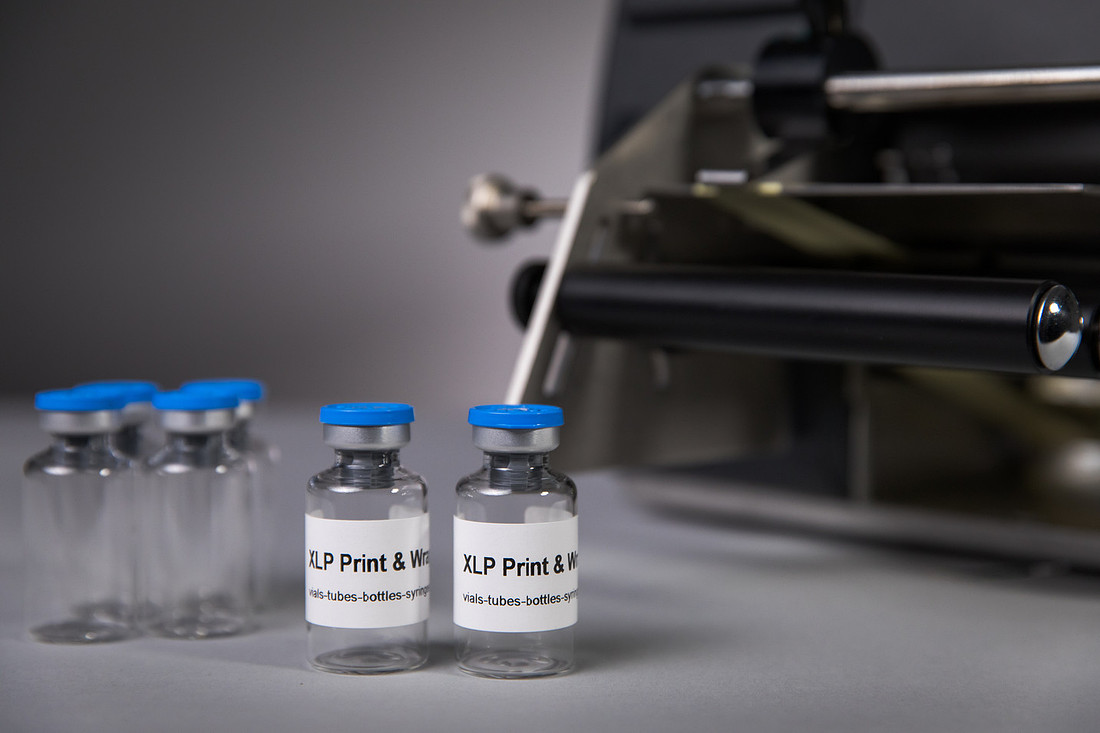 Two clear glass vials labelled "XLP Print & Wrap" in front of the black rollers of the machine.