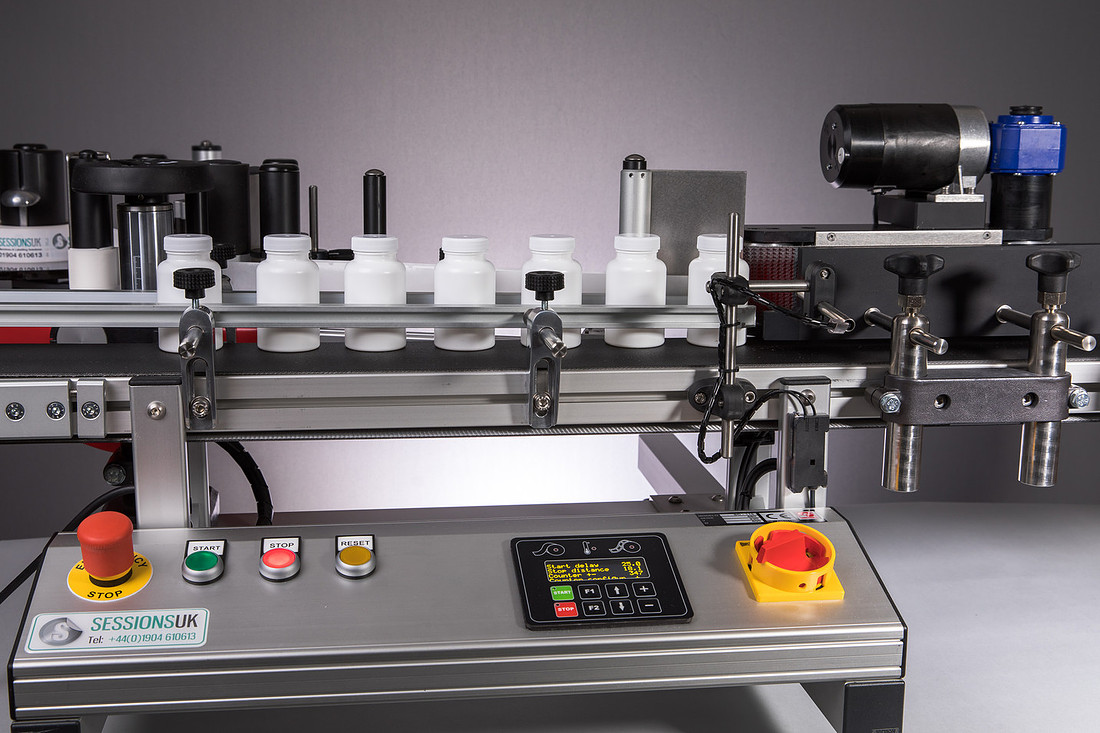 An inline labelling machine with white bottles along the conveyor and its push-button control panel in the foreground.